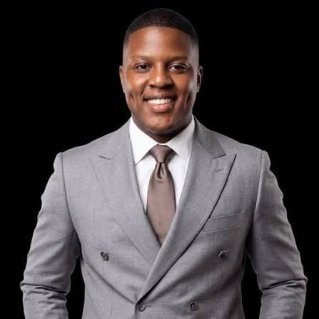 Ramel Newerls smiling in a gray double-breasted suit and brown tie against a black background.